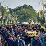 Large crowd of people in blue attire in a funeral or ceremonial procession, carrying a decorated bier under an arched gateway.