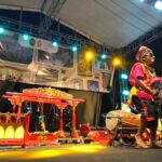 Performer in colorful traditional costume and hat on stage at night, holding a feathered prop fan during a cultural show with red ornate instruments nearby.