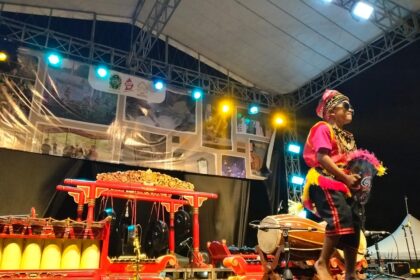 Performer in colorful traditional costume and hat on stage at night, holding a feathered prop fan during a cultural show with red ornate instruments nearby.