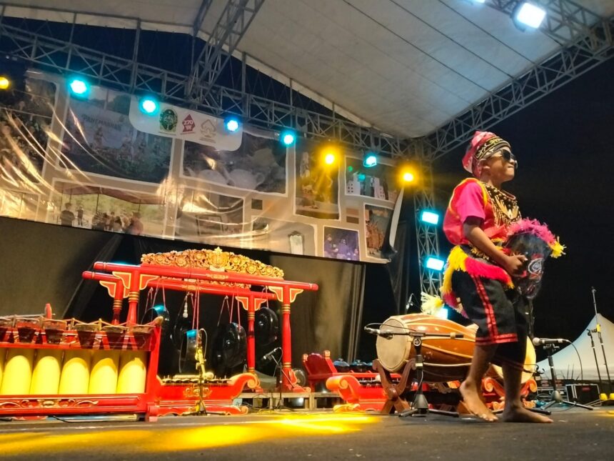 Performer in colorful traditional costume and hat on stage at night, holding a feathered prop fan during a cultural show with red ornate instruments nearby.