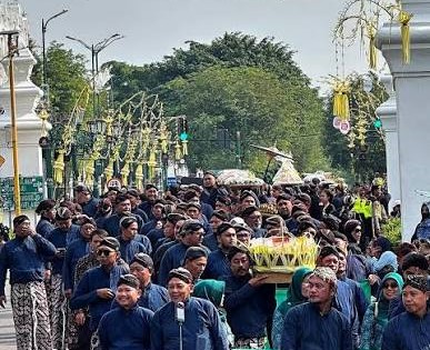 Large crowd of people in blue attire in a funeral or ceremonial procession, carrying a decorated bier under an arched gateway.