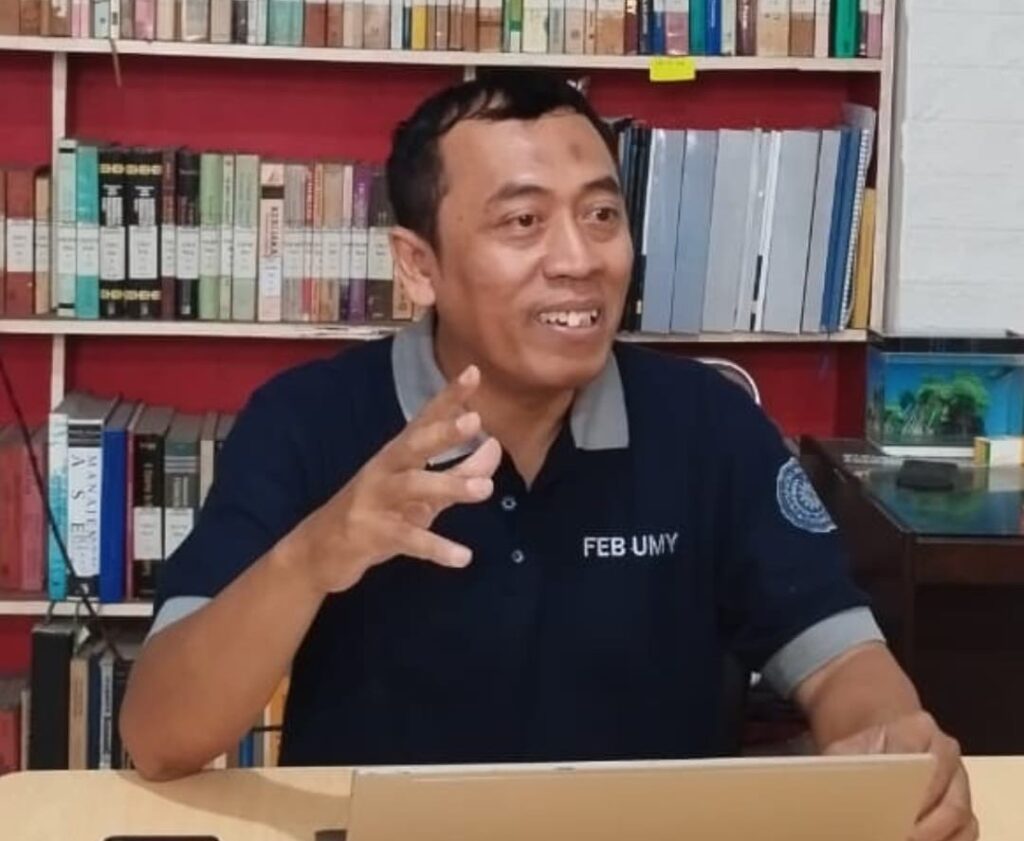 000 - Mabur.co Man in a navy polo with FEB UMY, gesturing while speaking in a library behind him with books on shelves.