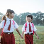 Two young schoolgirls in white shirts and red uniforms holding hands in a sunlit grassy field.
