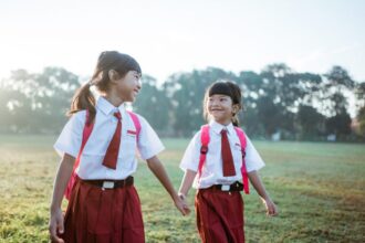 Two young schoolgirls in white shirts and red uniforms holding hands in a sunlit grassy field.
