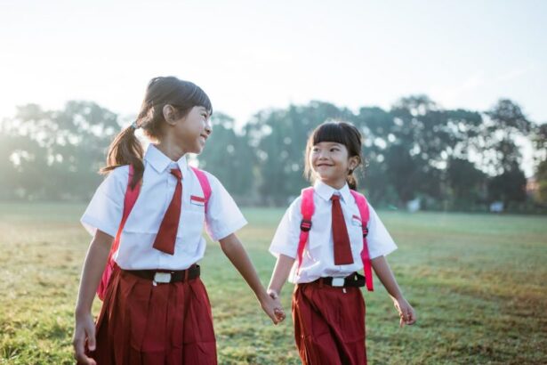 Two young schoolgirls in white shirts and red uniforms holding hands in a sunlit grassy field.