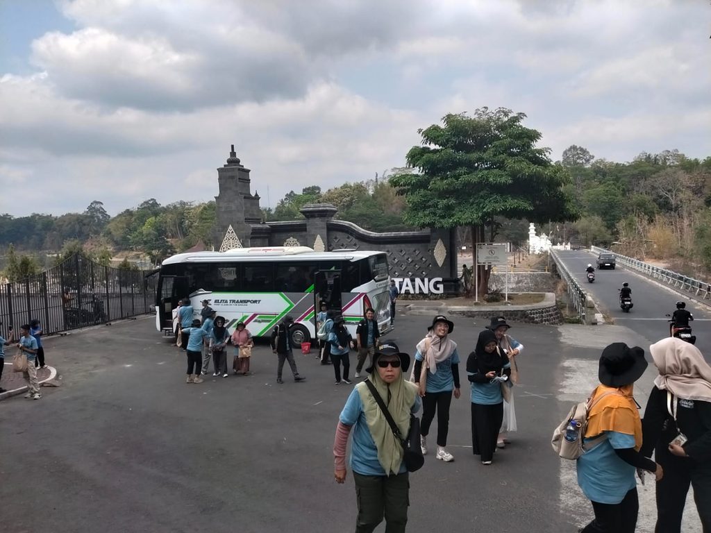 Tour group gathers near a white coach at a decorative stone entrance on a paved lot, cloudy sky above.