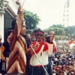 Speaker on a stage raises his arm into the air, addressing a large outdoor political rally with security nearby and a crowd watching intently.