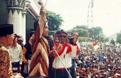 Speaker on a stage raises his arm into the air, addressing a large outdoor political rally with security nearby and a crowd watching intently.