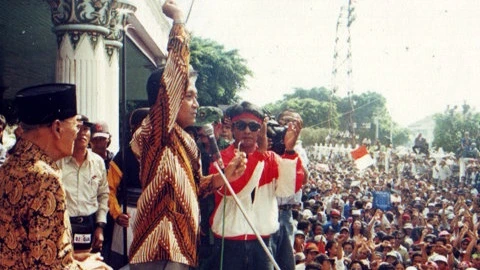 Speaker on a stage raises his arm into the air, addressing a large outdoor political rally with security nearby and a crowd watching intently.