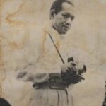 Man in vintage clothing holding a camera, posing for a photograph, sepia-toned portrait style.