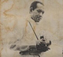 Man in vintage clothing holding a camera, posing for a photograph, sepia-toned portrait style.