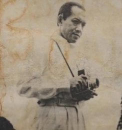 Man in vintage clothing holding a camera, posing for a photograph, sepia-toned portrait style.