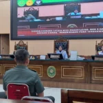 Courtroom with three judges at a raised bench, two uniformed men seated in the foreground, and a large monitor showing the hearing.