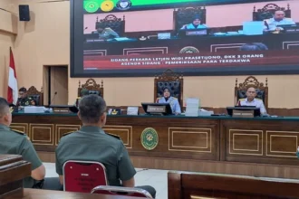 Courtroom with three judges at a raised bench, two uniformed men seated in the foreground, and a large monitor showing the hearing.