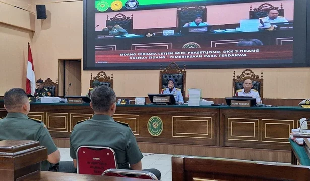Courtroom with three judges at a raised bench, two uniformed men seated in the foreground, and a large monitor showing the hearing.