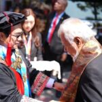 Graduate in cap and gown shaking hands with an older man wearing a decorative stole at an outdoor graduation ceremony, with onlookers in the background.