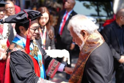 Graduate in cap and gown shaking hands with an older man wearing a decorative stole at an outdoor graduation ceremony, with onlookers in the background.