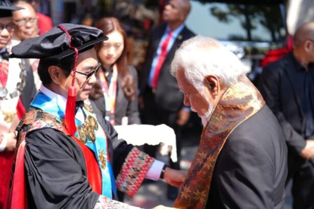 Graduate in cap and gown shaking hands with an older man wearing a decorative stole at an outdoor graduation ceremony, with onlookers in the background.