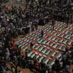 Large crowd gathers around dozens of caskets arranged in a grid, each draped with green, white, and red flags at a funeral ceremony.