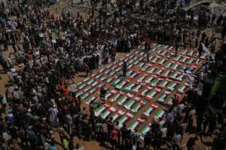 Large crowd gathers around dozens of caskets arranged in a grid, each draped with green, white, and red flags at a funeral ceremony.