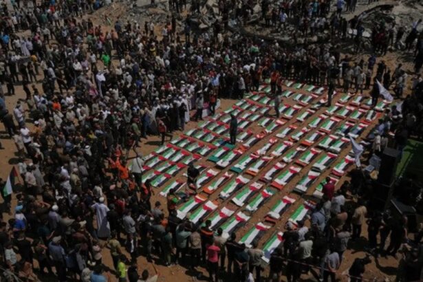 Large crowd gathers around dozens of caskets arranged in a grid, each draped with green, white, and red flags at a funeral ceremony.
