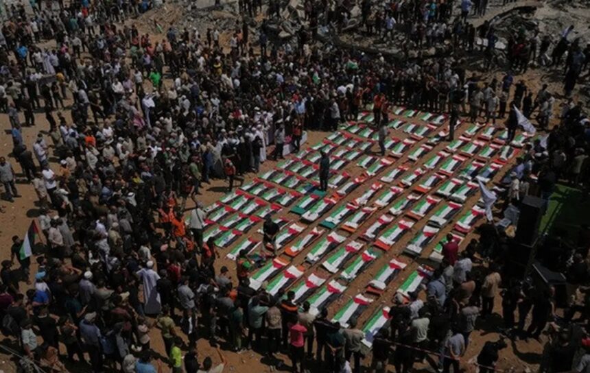 Large crowd gathers around dozens of caskets arranged in a grid, each draped with green, white, and red flags at a funeral ceremony.