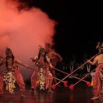 Group of dancers in traditional red patterned outfits with headdresses, performing with long spears on a dim stage with red smoke in the background for a cultural performance.