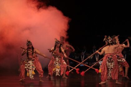 Group of dancers in traditional red patterned outfits with headdresses, performing with long spears on a dim stage with red smoke in the background for a cultural performance.