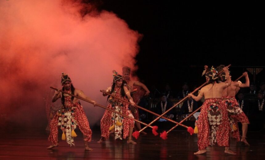 Group of dancers in traditional red patterned outfits with headdresses, performing with long spears on a dim stage with red smoke in the background for a cultural performance.