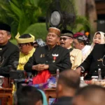 Group of men in black traditional outfits and headwear seated at a ceremonial outdoor event, with medals and drinks on the table