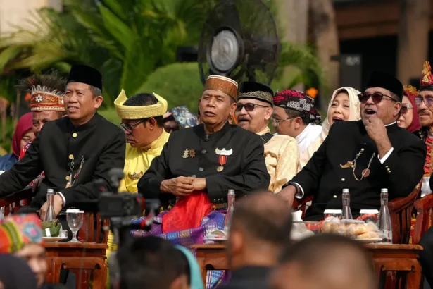 Group of men in black traditional outfits and headwear seated at a ceremonial outdoor event, with medals and drinks on the table