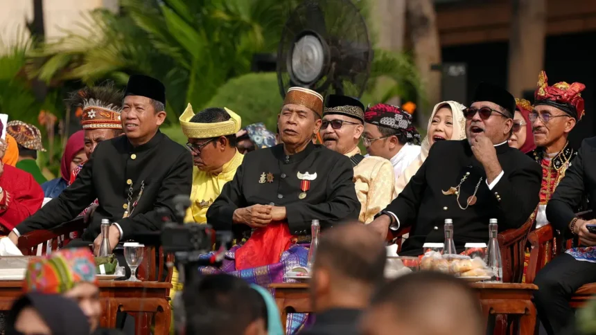 Group of men in black traditional outfits and headwear seated at a ceremonial outdoor event, with medals and drinks on the table