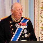 Older man in a dark ceremonial military uniform with gold epaulettes and medals at a podium in a richly decorated room, speaking.