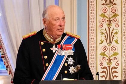 Older man in a dark ceremonial military uniform with gold epaulettes and medals at a podium in a richly decorated room, speaking.