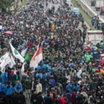 Massive protest on a city highway with hundreds of protesters marching and waving red-and-white flags.