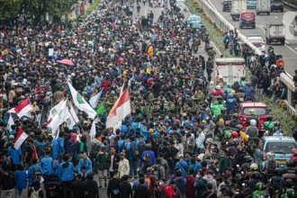 Massive protest on a city highway with hundreds of protesters marching and waving red-and-white flags.