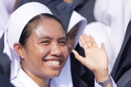 Smiling woman in a white nurse's uniform and headscarf waves at the camera.