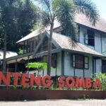 Two-story building with a brown roof behind tall palm trees, and a large red sign in front reading 'Benteng Somba Opu'