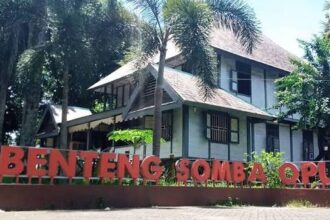 Two-story building with a brown roof behind tall palm trees, and a large red sign in front reading 'Benteng Somba Opu'