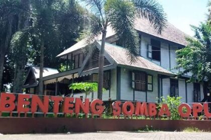 Two-story building with a brown roof behind tall palm trees, and a large red sign in front reading 'Benteng Somba Opu'