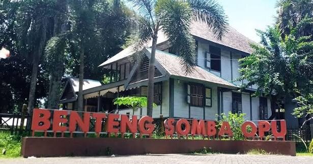 Two-story building with a brown roof behind tall palm trees, and a large red sign in front reading 'Benteng Somba Opu'