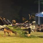 Nighttime festival scene with several performers running on a field, pulling a long palm-frond rope as part of a cultural act.