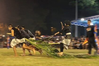 Nighttime festival scene with several performers running on a field, pulling a long palm-frond rope as part of a cultural act.