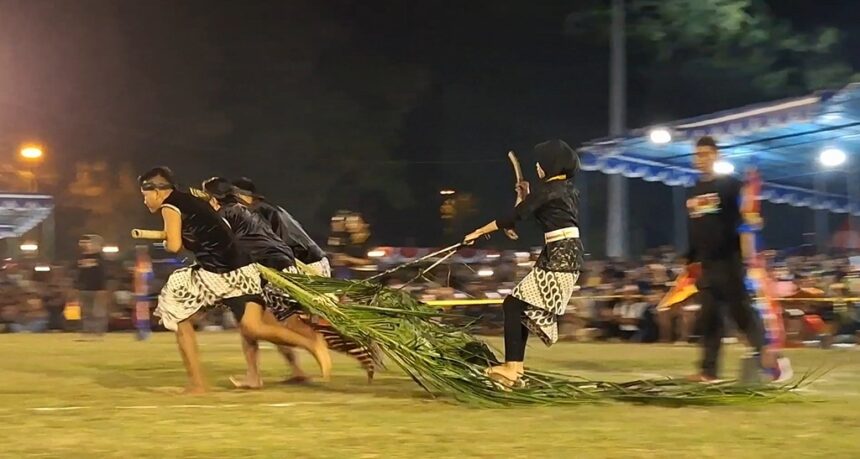 Nighttime festival scene with several performers running on a field, pulling a long palm-frond rope as part of a cultural act.
