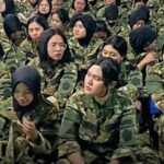 A group of female soldiers in camouflage uniforms seated closely together, many wearing hijabs.