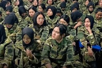 A group of female soldiers in camouflage uniforms seated closely together, many wearing hijabs.