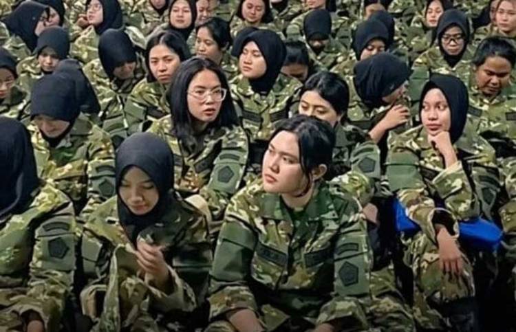 A group of female soldiers in camouflage uniforms seated closely together, many wearing hijabs.