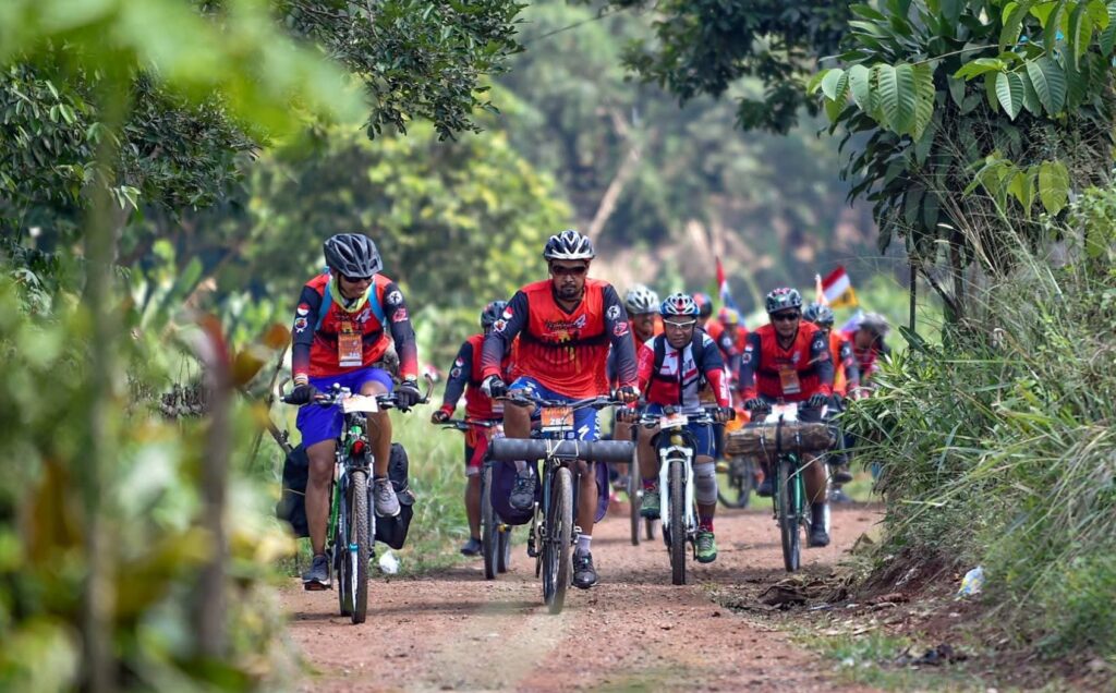 1000265005 - Mabur.co Group of cyclists riding on a dirt path through dense green foliage, front riders in red jerseys and helmets during a race.