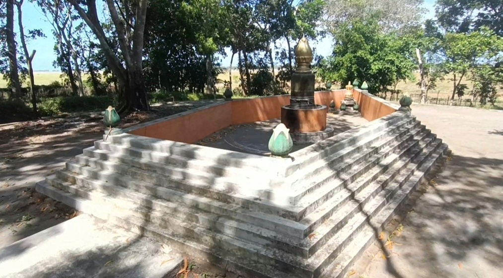 Concrete stepped platform surrounding a central small shrine with a golden stupa in a sunlit outdoor area. Trees and shadows frame the scene.
