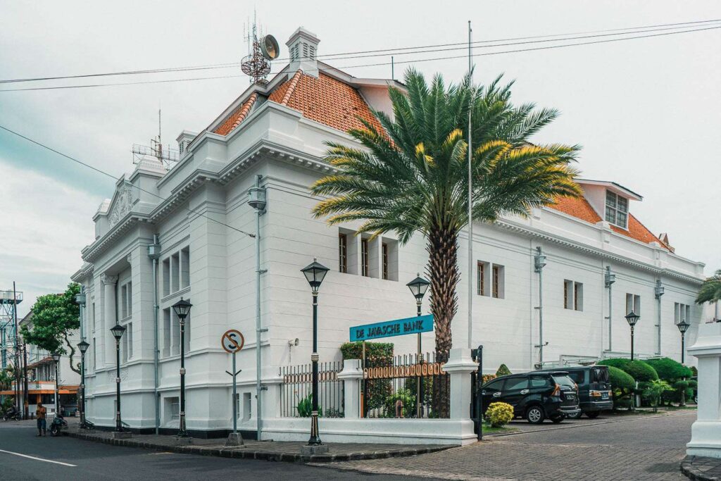1000282525 - Mabur.co White historic bank building with orange-tiled roof on a street corner, palm tree in front and iron gate signage reading'De Javaasche Bank'